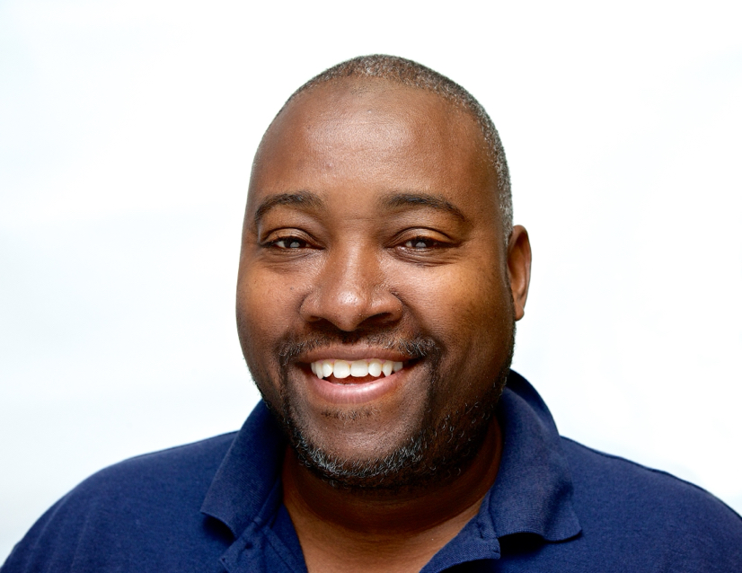 A professional headshot os a smiling man with short grey hair and a beard, wearing a navy blue shirt, against a white background.