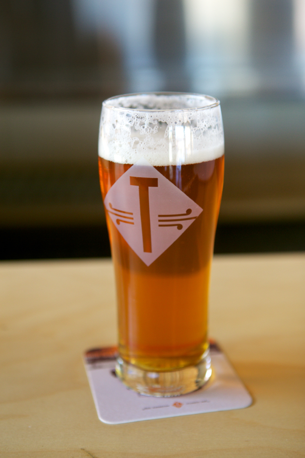 A glass of amber Temperance beer with a logo on the side, sitting on a coaster on a wooden table.